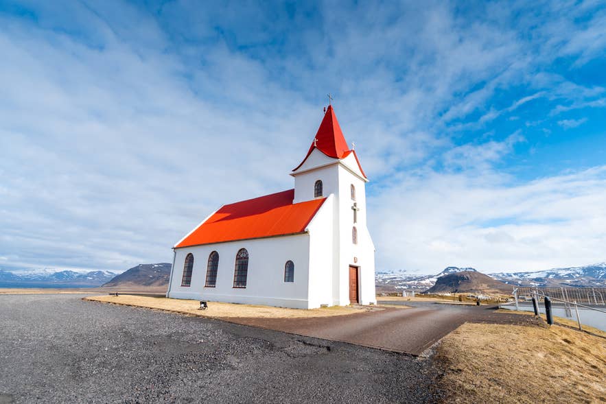 Ingjaldsholskirkja Church, white with a red roof, under blue sky with mountains behind in Snaefellsnes Peninsula