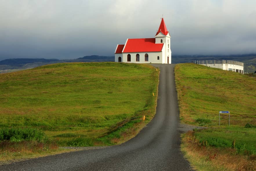 A straight road leading up a green hill to Ingjaldsholskirkja Church under a cloudy sky