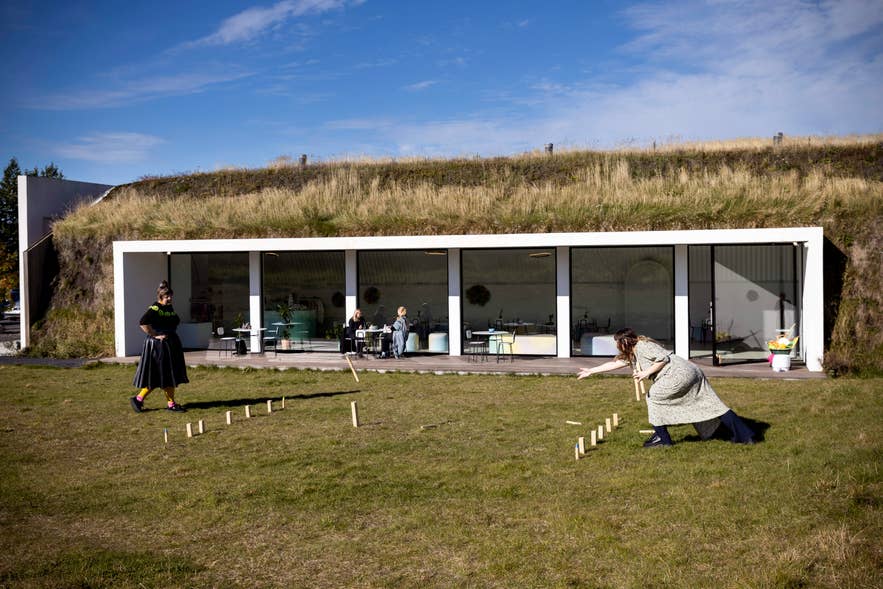 Visitors playing a lawn game outside Hofudstodin, with the building's grass-covered roof and glass-walled cafe behind.
