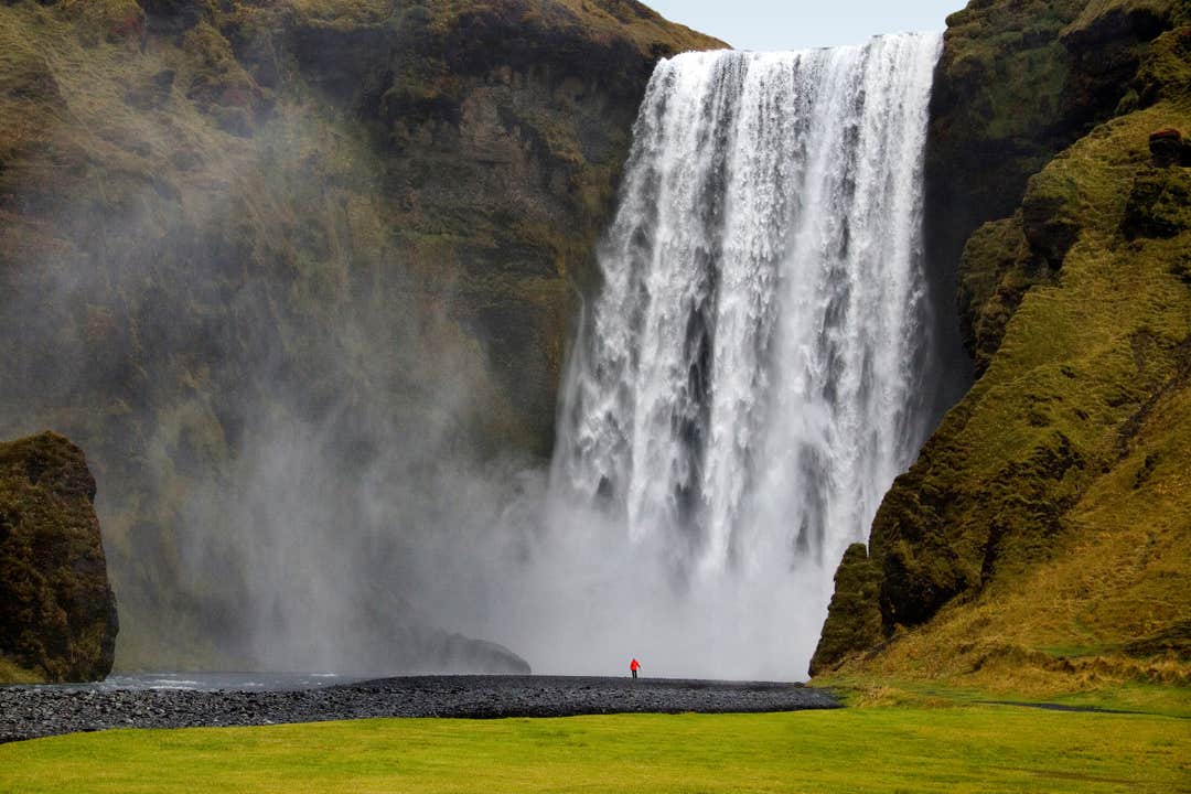 Skogafoss vattenfall som störtar ner från branta klippor, med dimma som stiger upp över det gröna gräset i förgrunden.