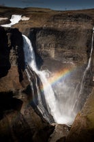 A rainbow shines in front of a picturesque Icelandic waterfall amid rugged scenery.