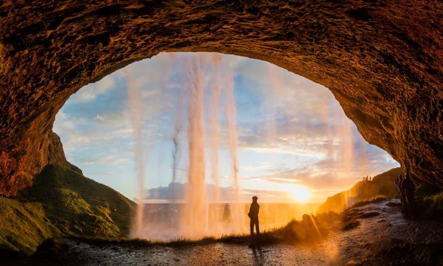 A visitor standing inside the cave behind Seljalandsfoss waterfall as the midnight sun blazes through the falling water.
