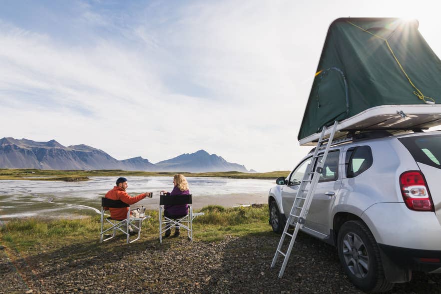 Ett par kopplar av i campingstolar bredvid en fyrhjulsdriven bil med taktält, med utsikt över berg och vatten under en ljus sommardag på Island.