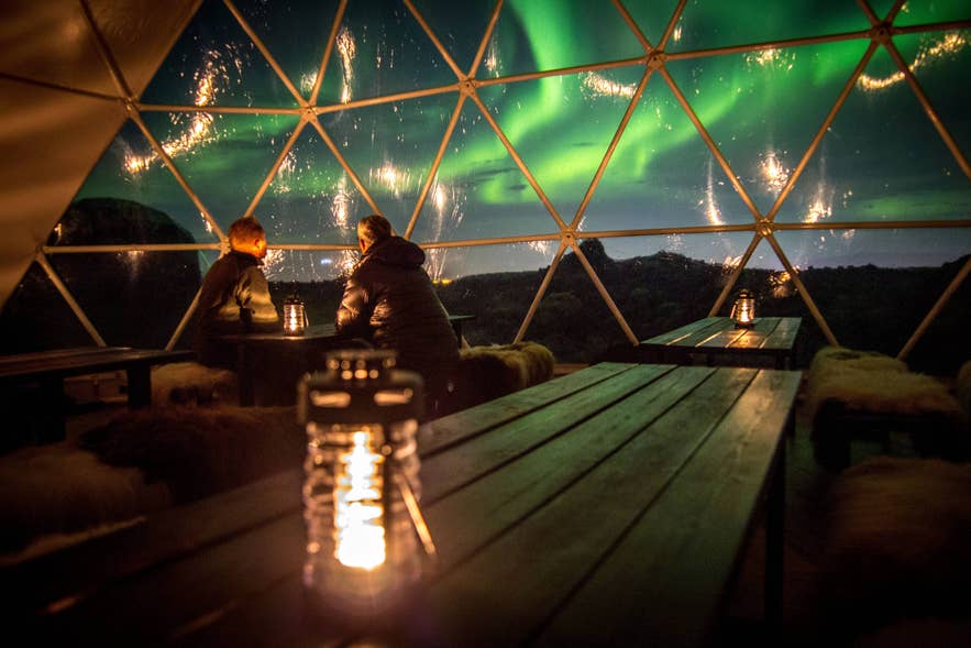 Two people in a glass dome watching green northern lights, with a lantern on the table