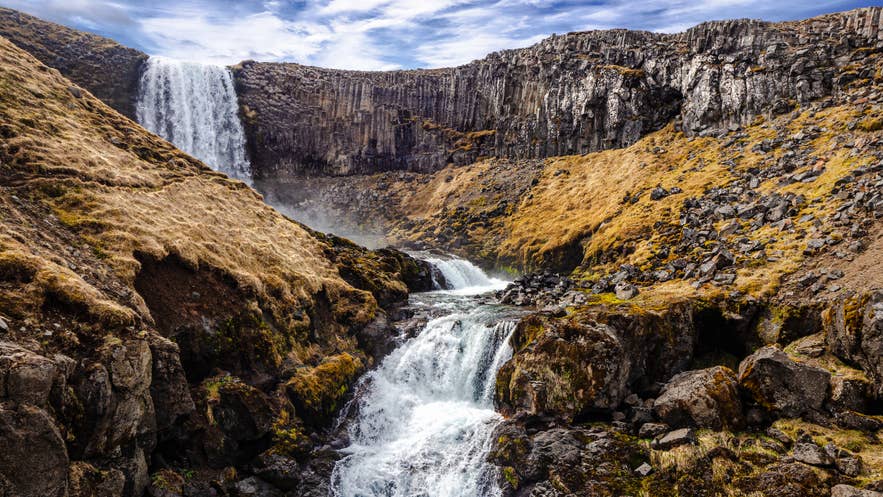 Svodufoss Waterfall and its lower cascade framed by basalt cliffs and golden hillsides.