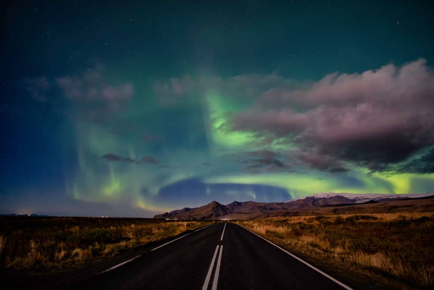 Northern lights glow above an empty road in Iceland, with green auroras, clouds, stars, and dark mountains in the distance.