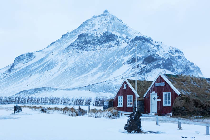 Red turf-roofed houses in Arnarstapi under a snow-covered mountain in winter.