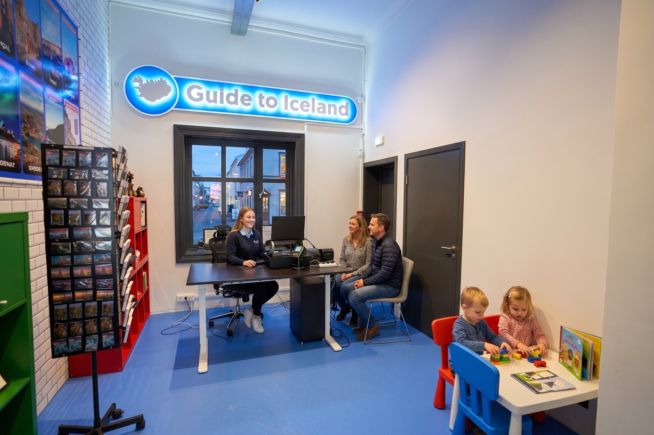 Recepcjonistka pomagająca dwóm podróżnym przy punkcie informacji turystycznej Guide to Iceland, obok bawią się dzieci.