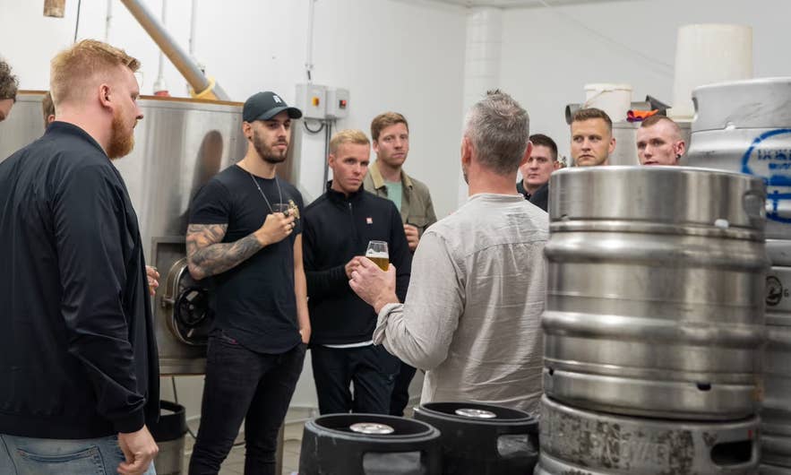 A group on a brewery tour holding beers beside steel tanks and kegs