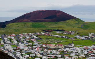 The red scoria cone of Eldfell Volcano rises above green fields in Vestmannaeyjar.