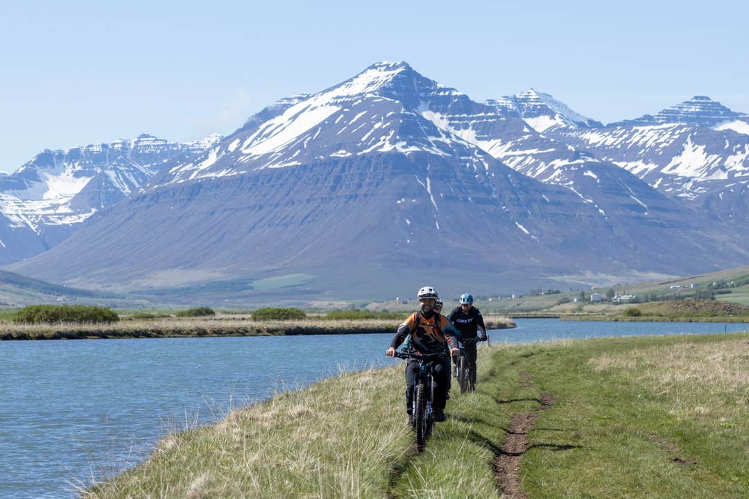 Two cyclists ride along a lakeside trail beneath snow-capped peaks in Svarfadardalur Valley near Dalvik.