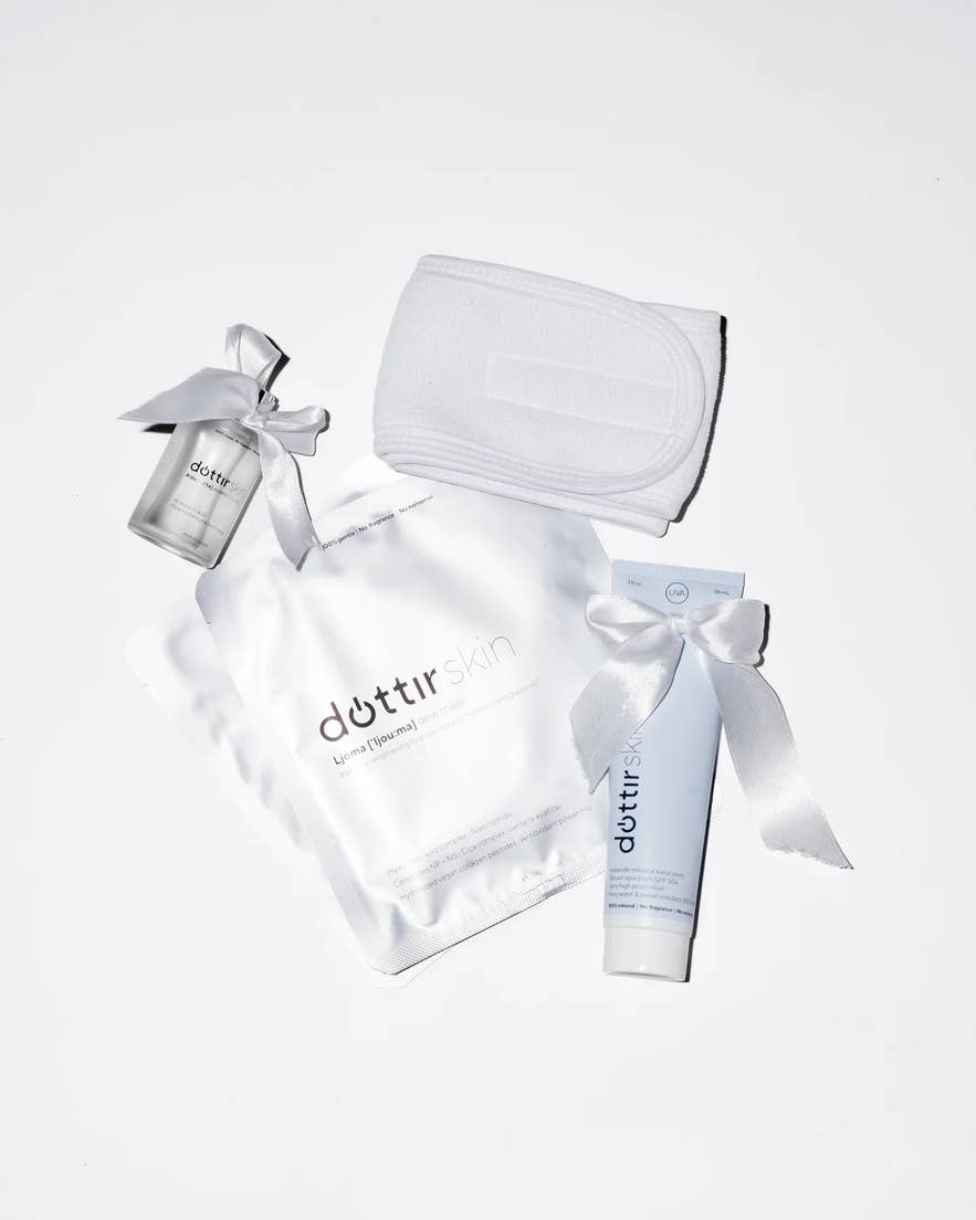 Flat-lay product photo of a Dóttir Skin skincare set on a white background, featuring a silver face-mask pouch, a white sunscreen tube, a small serum bottle tied with a satin bow, and a white spa headband.