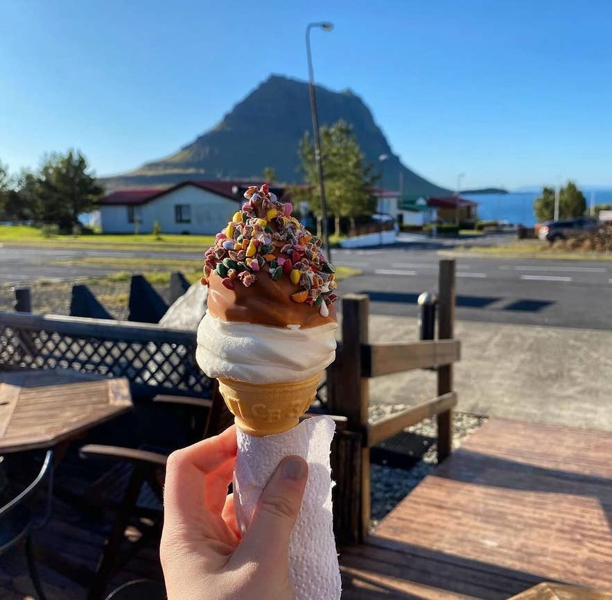 A soft-serve ice cream cone held up on the Kaffi 59 terrace, with Kirkjufell in the background.