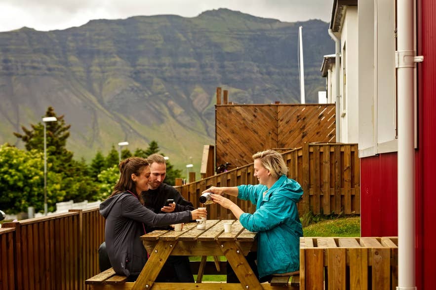 Guests share a picnic table outside red-roofed cabins in Grundarfjordur, below a steep mountainside.