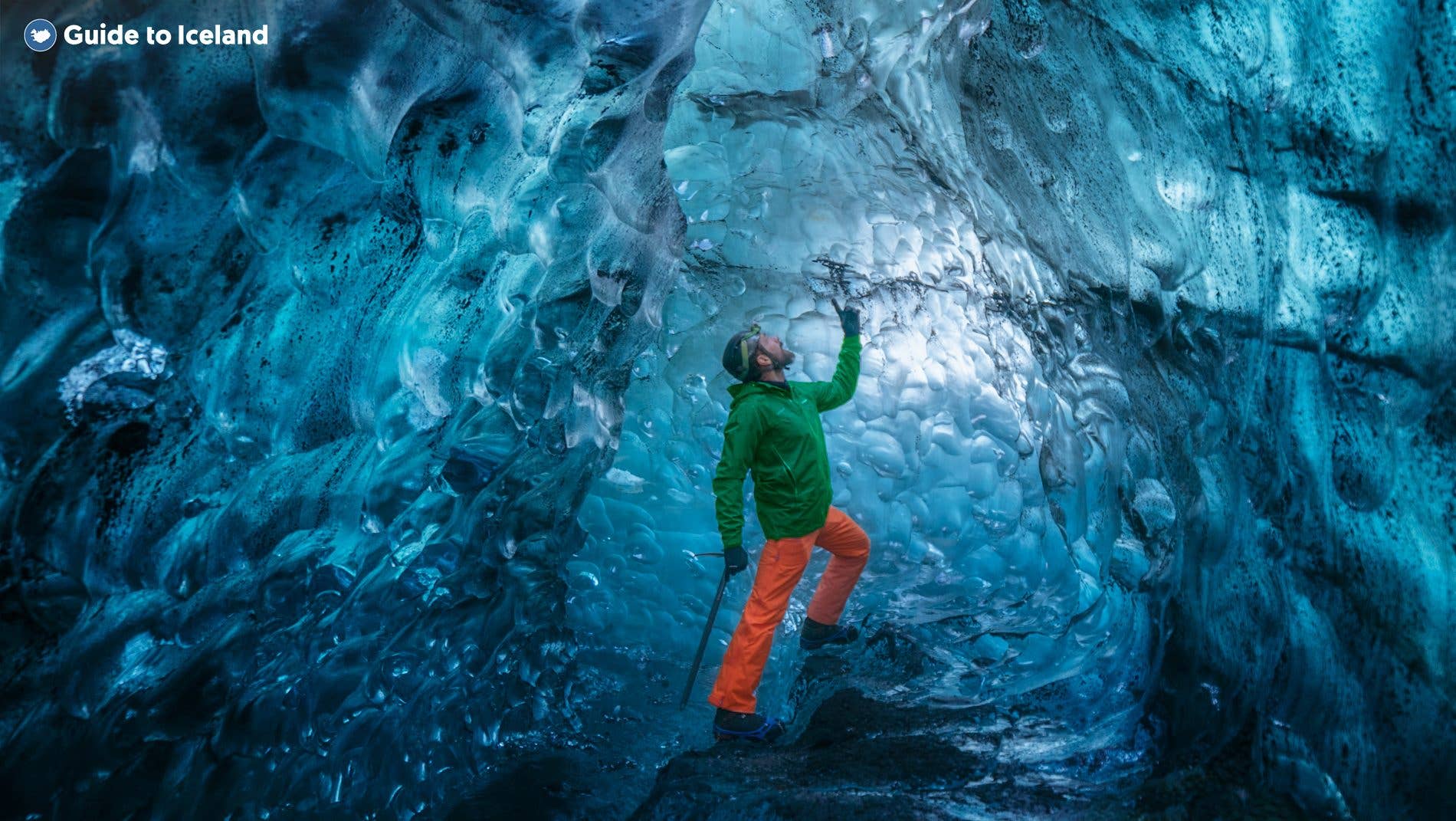 A visitor in a green jacket and orange trousers explores the deep-blue interior of a natural ice cave in Breiðamerkurjökull glac