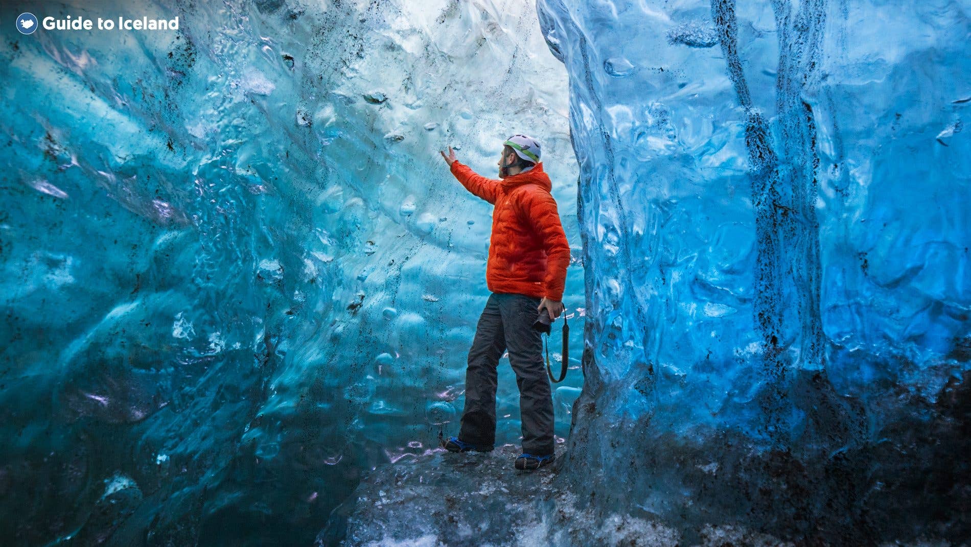 A person in a red jacket reaches toward the smooth blue wall of a natural glacier ice cave in Vatnajökull, southeast Iceland.