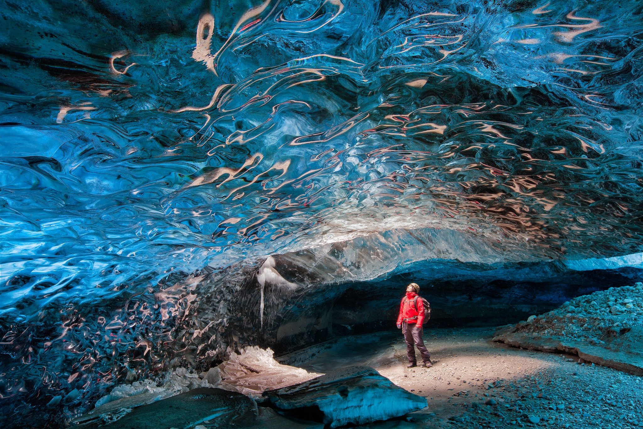 A lone visitor in red stands in a vast ice cave with a marbled blue-and-amber ceiling beneath Vatnajökull glacier, Iceland.