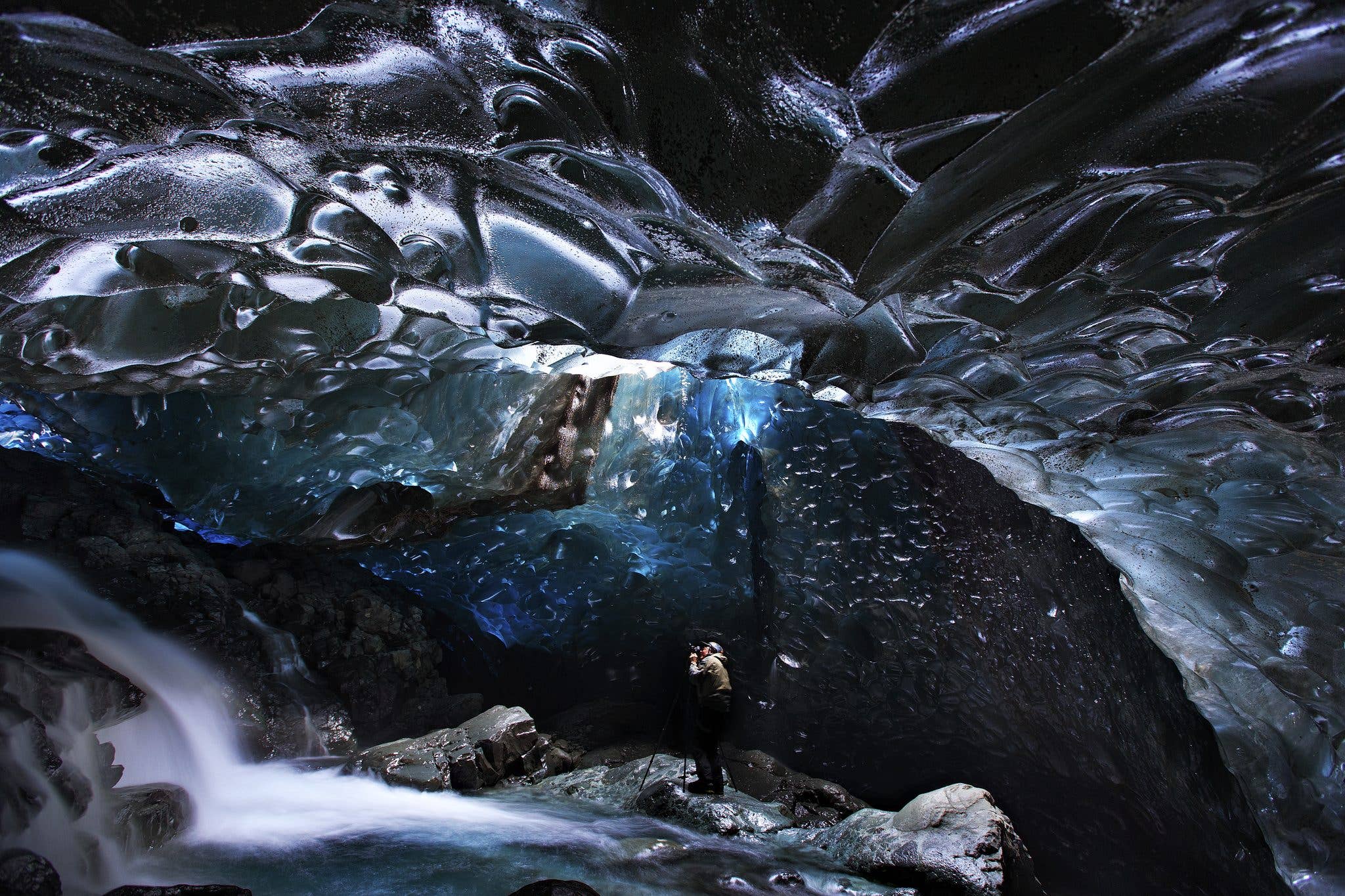 A photographer stands beside a meltwater stream inside a dark blue ice cave under Vatnajökull glacier, Iceland.