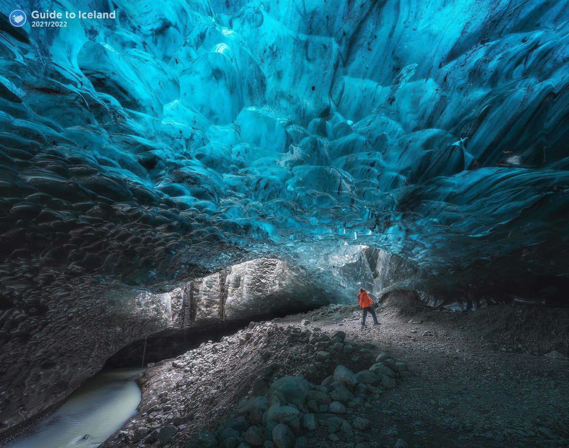 A traveller in red stands on the rocky floor of a brilliant blue ice cave under Vatnajökull glacier, Iceland.