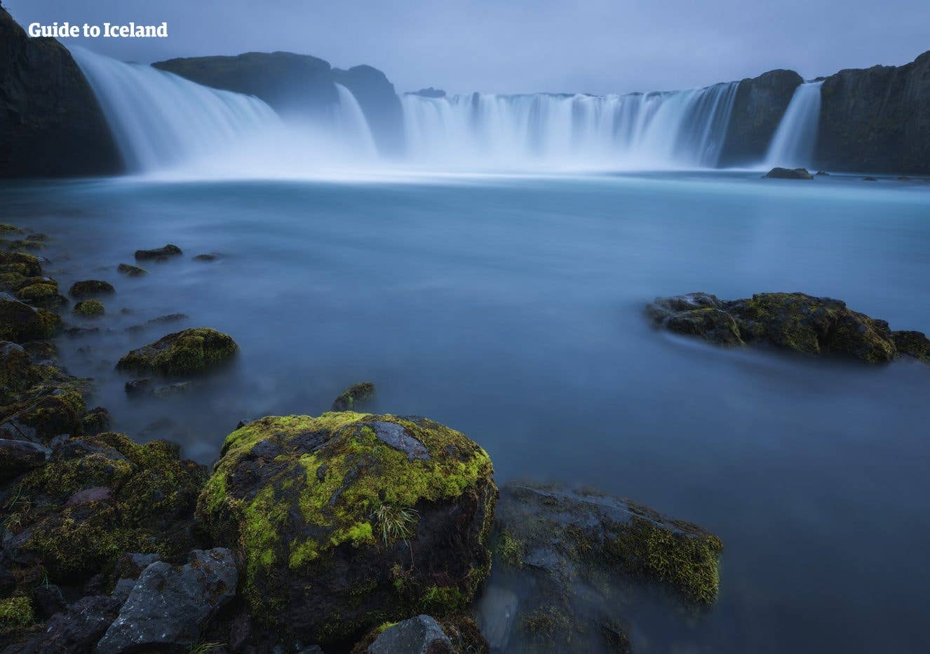 The wide, curved cascade of Goðafoss tumbles over moss-covered rocks on a summer evening in North Iceland.