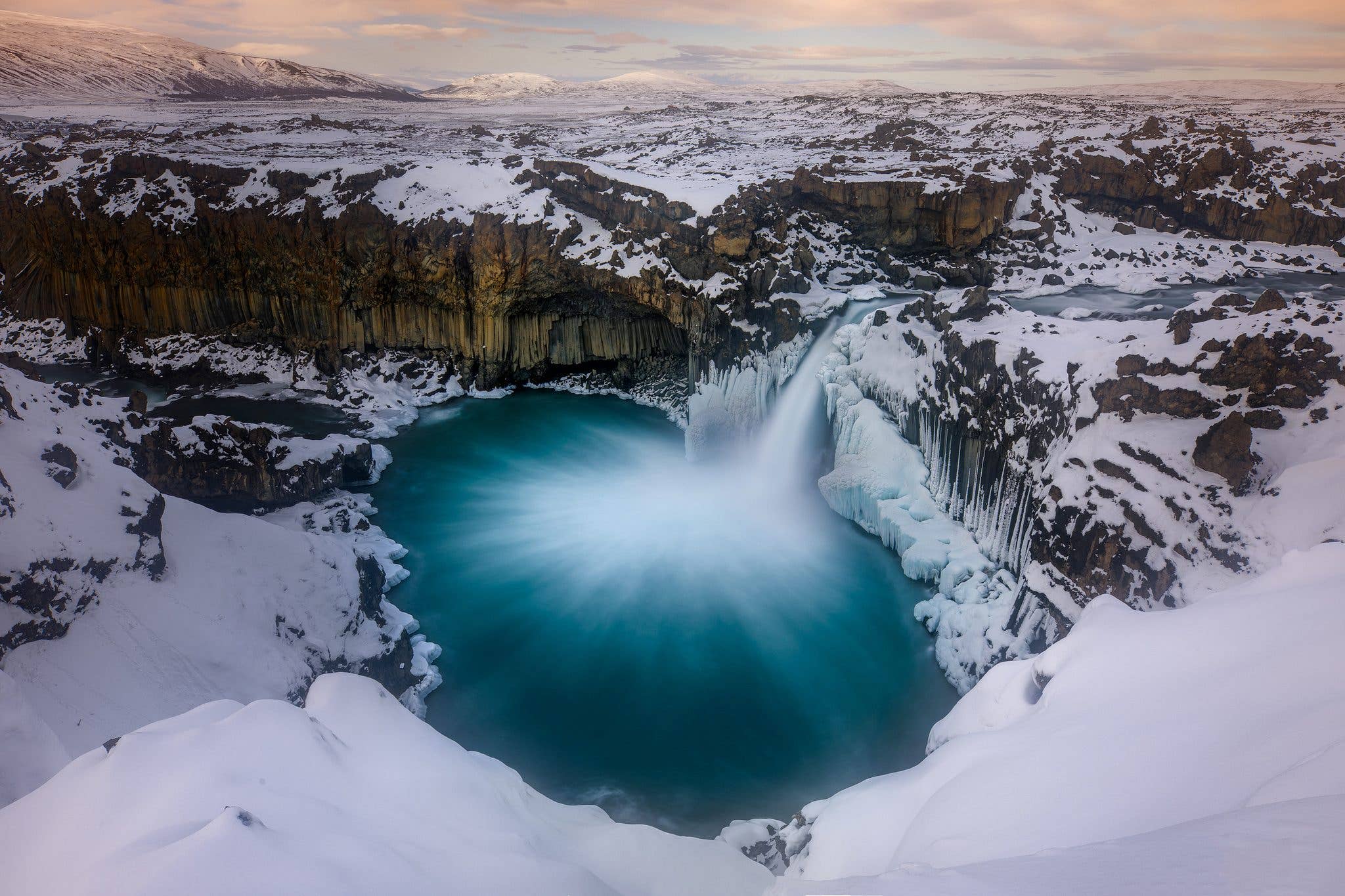 An aerial view of Aldeyjarfoss plunging into a turquoise pool ringed by basalt columns in the Icelandic Highlands.