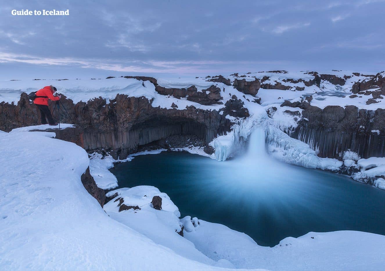 A photographer stands at the snow-covered edge of Aldeyjarfoss in the Highlands of North Iceland in winter.
