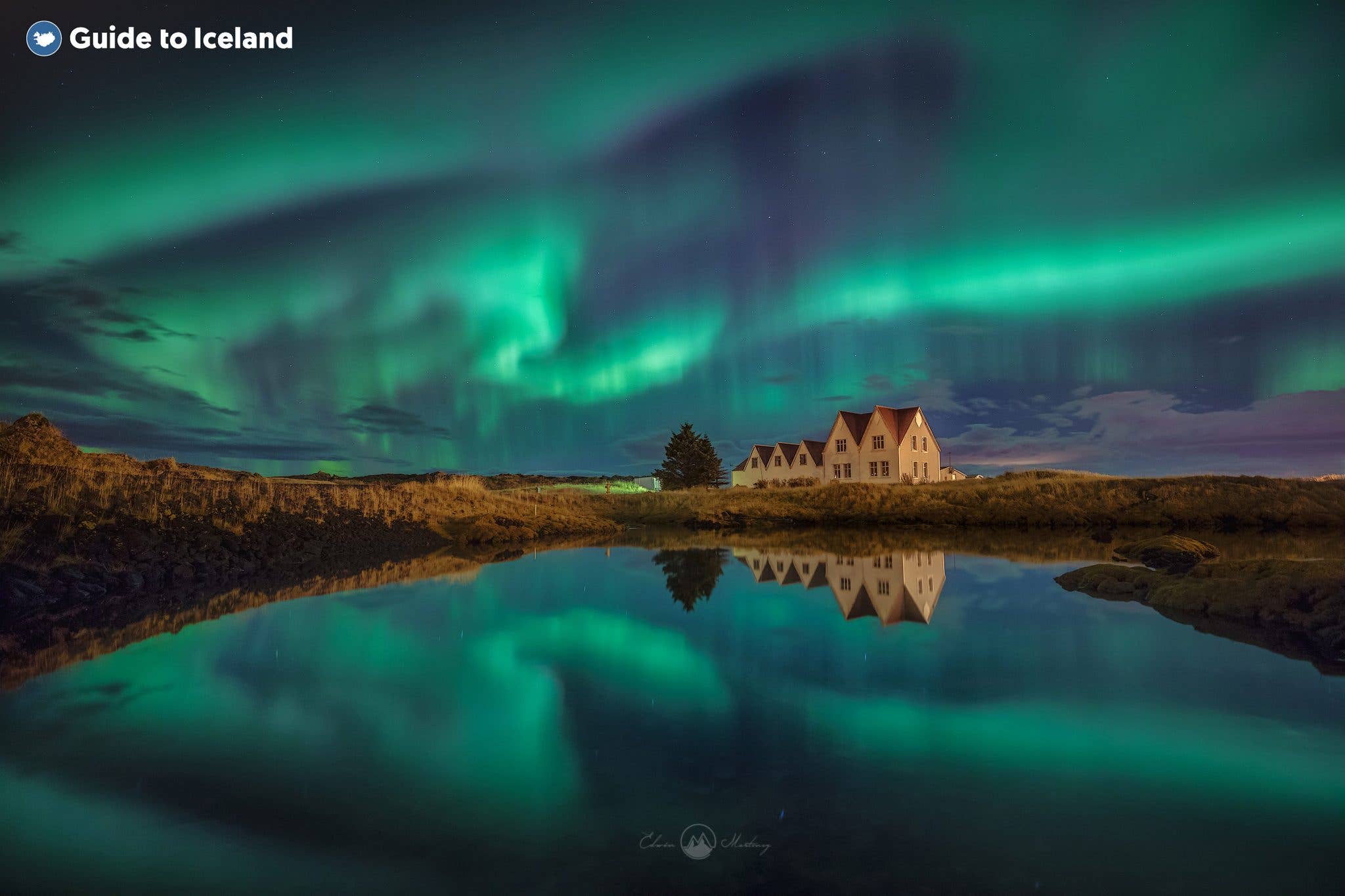 Vivid green aurora reflects in calm water beside traditional houses at Þingvellir, southwest Iceland.