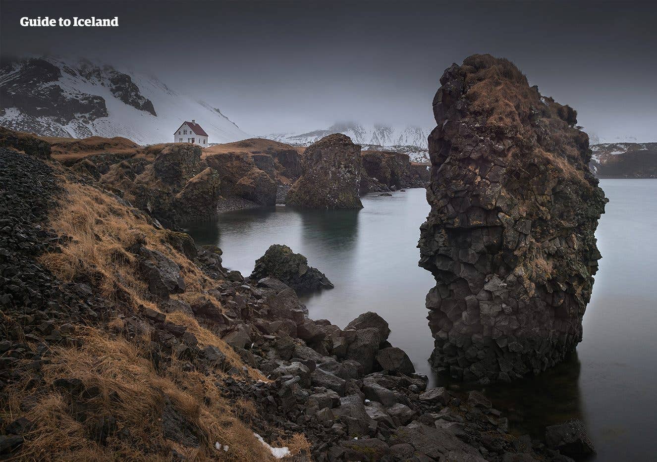 A tall sea stack rises off the coast at Arnarstapi, with a lone house behind, on the Snæfellsnes Peninsula, West Iceland.