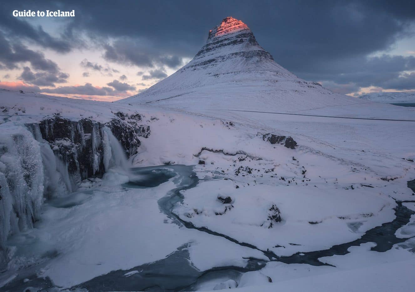 A snow-covered Kirkjufell glows at first light above Kirkjufellsfoss on the Snæfellsnes Peninsula, West Iceland.