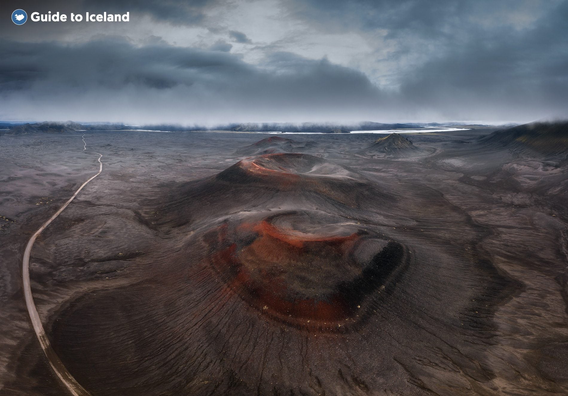 A red-rimmed volcanic crater cone sits amid the dark ash plains of the Icelandic Highlands, seen from above.