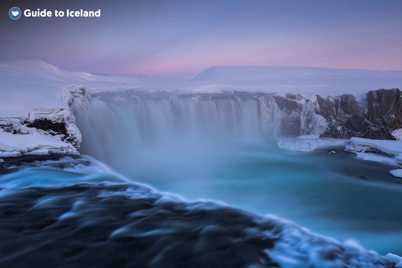 A soft pink winter sky glows above the snow-framed Goðafoss waterfall in North Iceland.