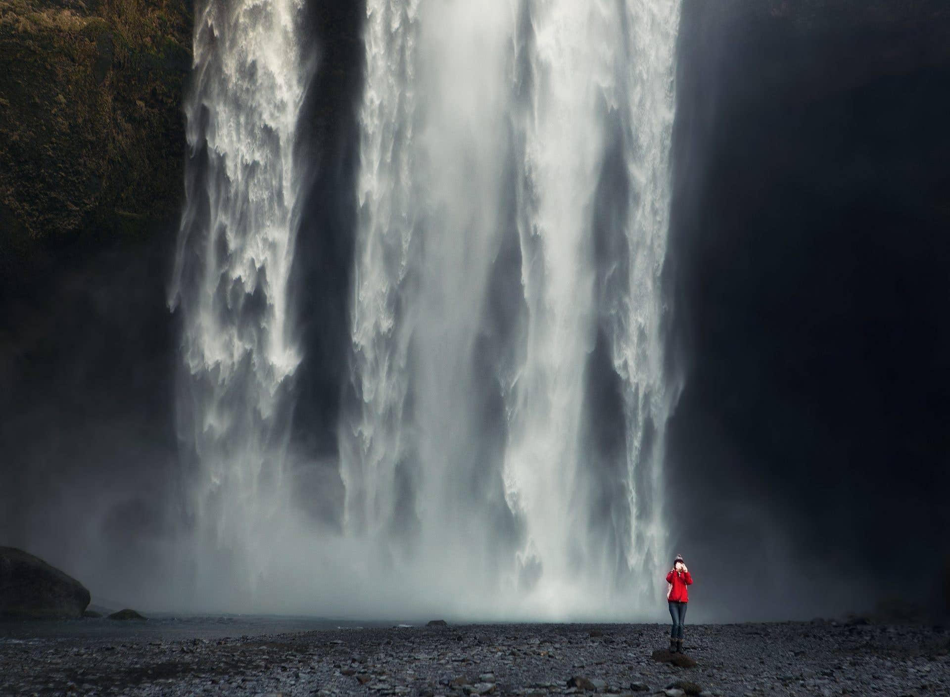 A visitor in red stands before the wide, powerful curtain of Skógafoss waterfall on the south coast of Iceland.