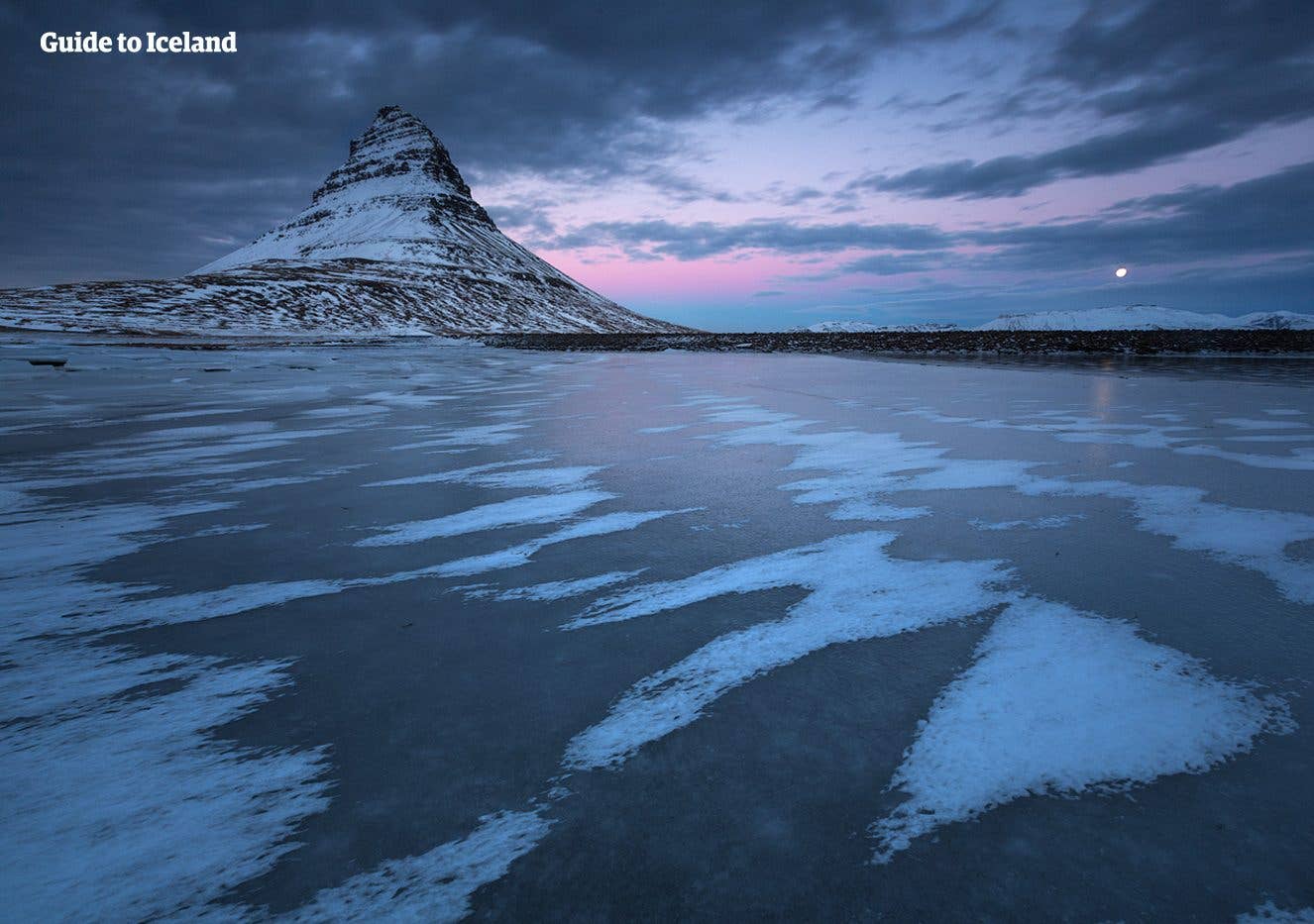 Kirkjufell mountain stands beyond a partly frozen shore under a soft winter sky on the Snæfellsnes Peninsula, Iceland.