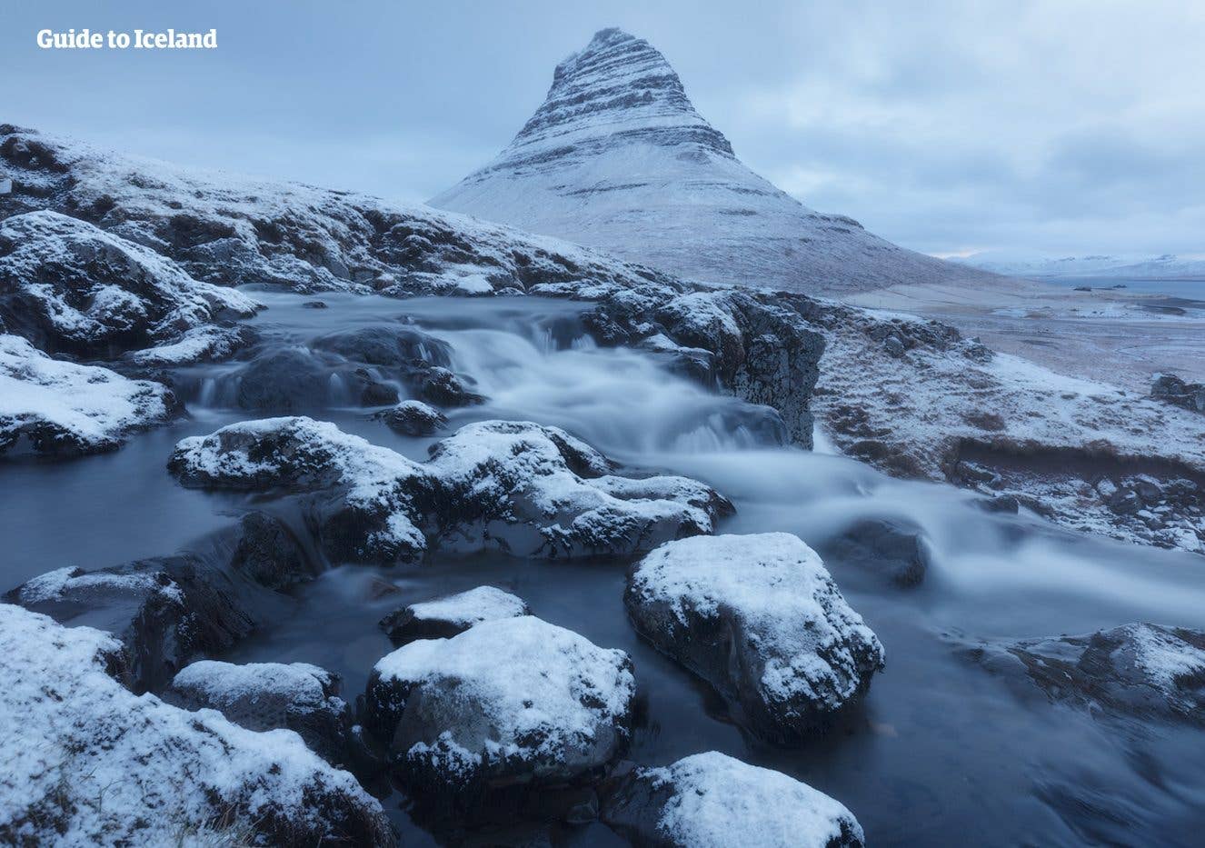 Snow-dusted boulders lead toward Kirkjufell mountain on a winter day on the Snæfellsnes Peninsula, Iceland.