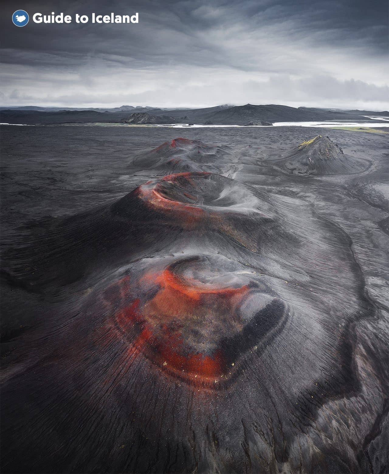Twin volcanic craters with red rims sit on black ash plains in the Icelandic Highlands, seen from the air.