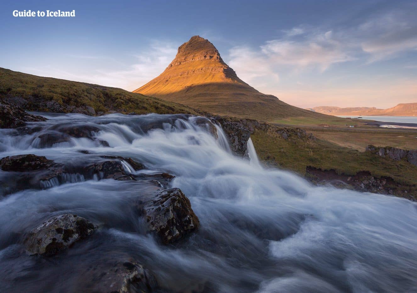 Water rushes over Kirkjufellsfoss below the pointed Kirkjufell mountain at sunset on the Snæfellsnes Peninsula, Iceland.
