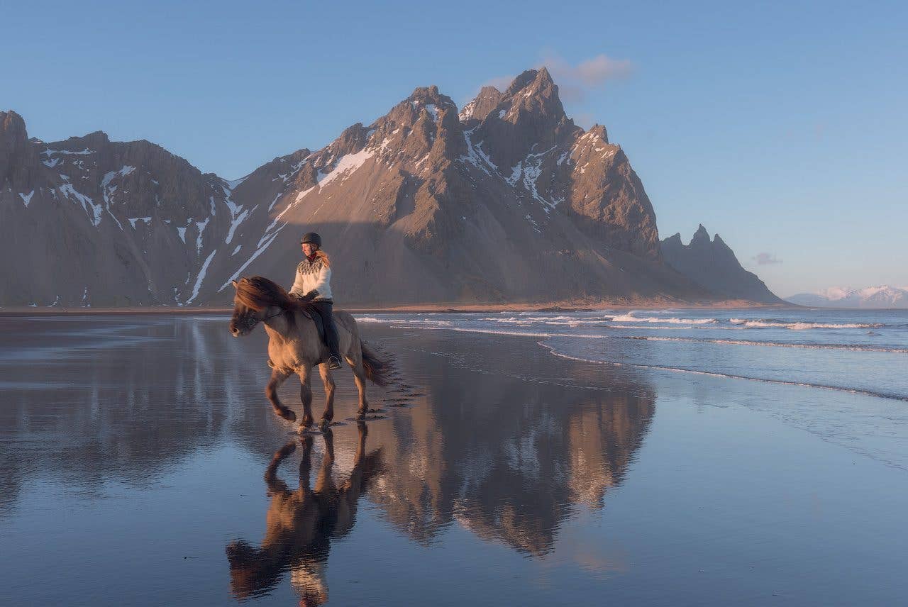 A rider on an Icelandic horse crosses a mirror-still beach beneath the Eystrahorn mountains in southeast Iceland.