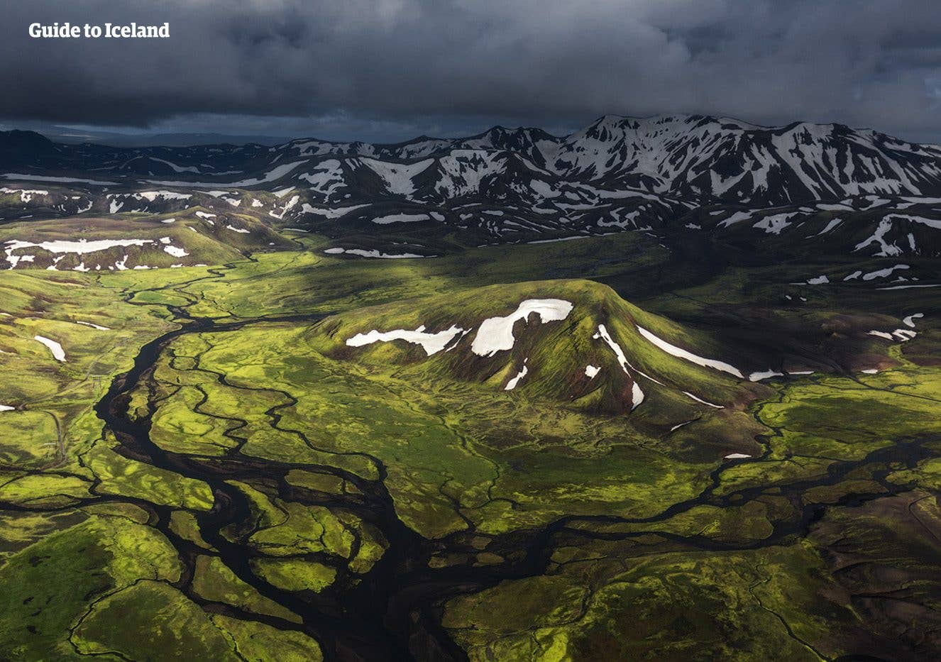 An aerial view of a braided river winding between green rhyolite mountains in the Icelandic Highlands.
