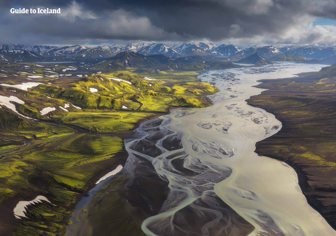 A glacial river fans across the plains below green Highland mountains, seen from above in Iceland.