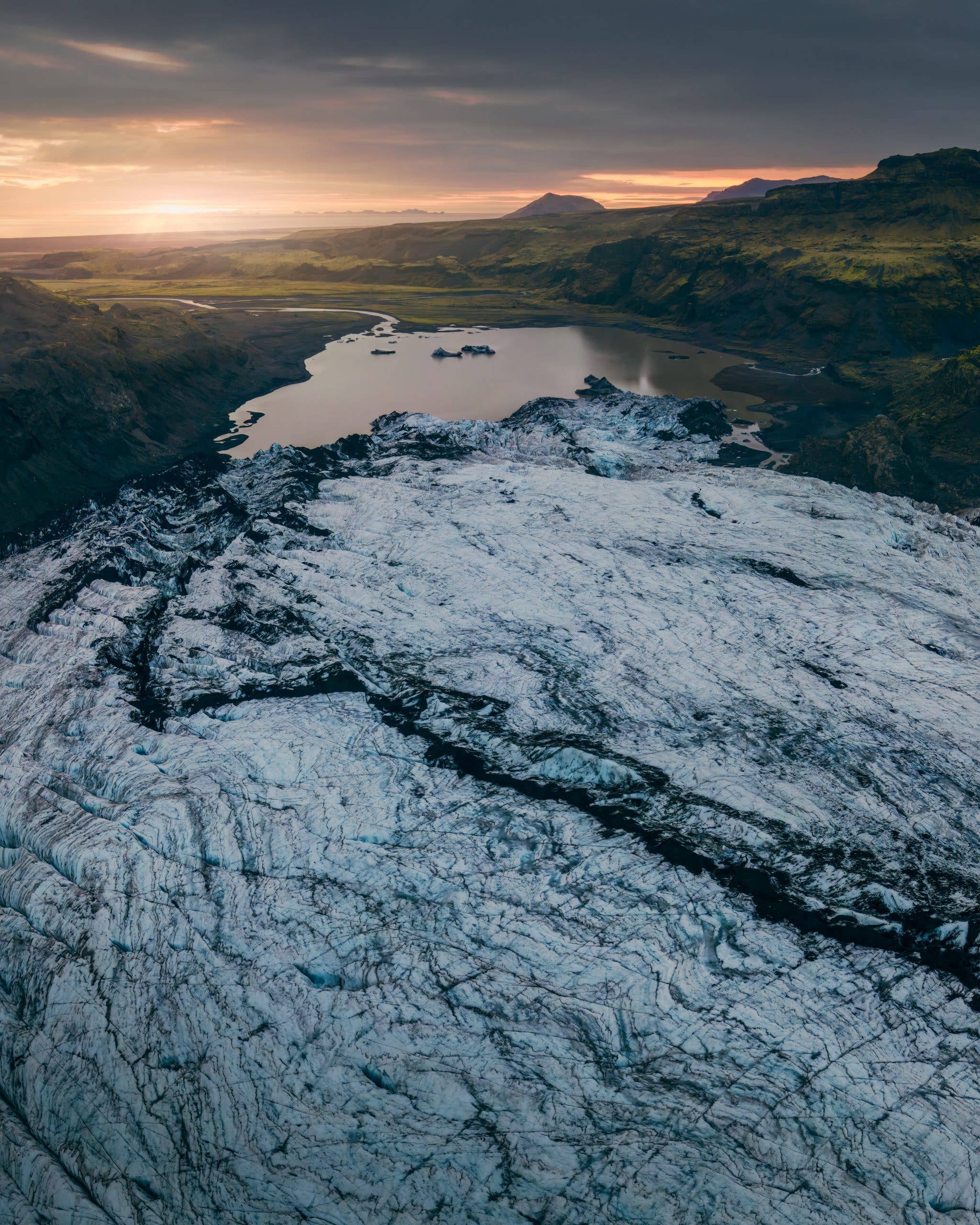 An aerial view of a glacier tongue meeting green mountains at sunrise in Iceland.