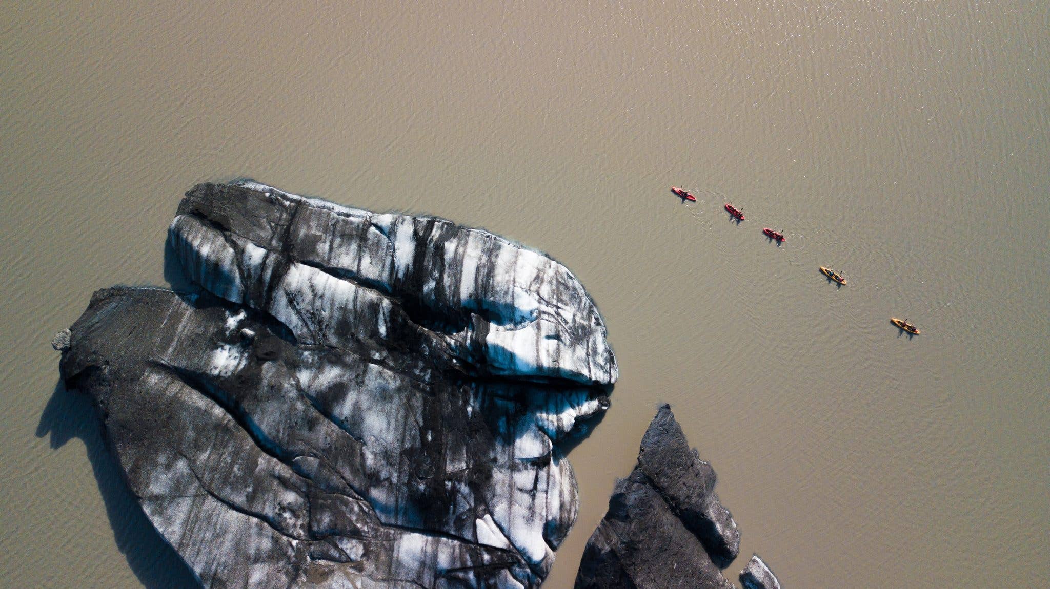 A kayaker glides across a calm glacial lagoon ringed by ice, seen from the air in Iceland.
