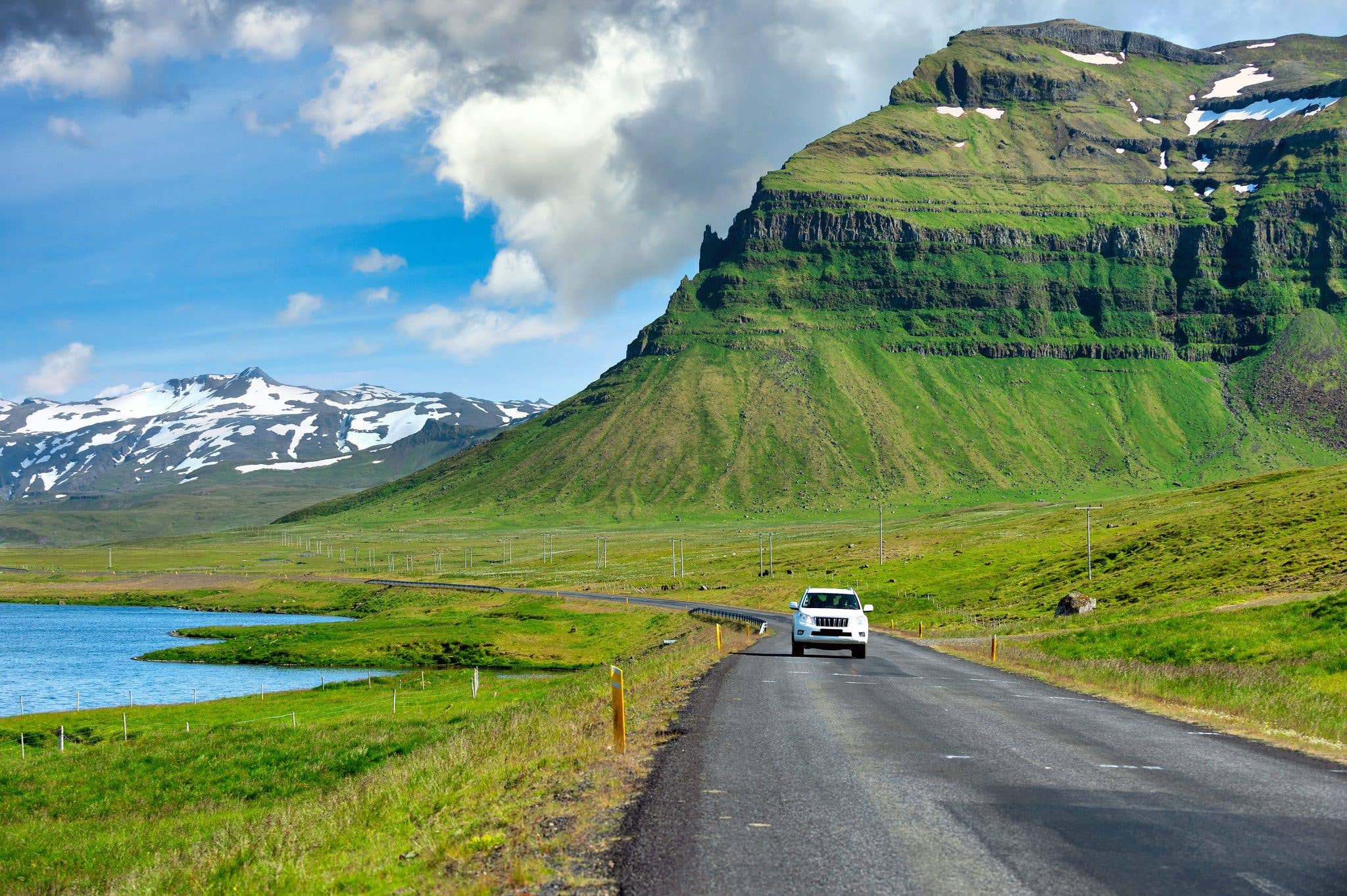 A car follows an empty road past a green fjordside mountain in the Westfjords of Iceland.