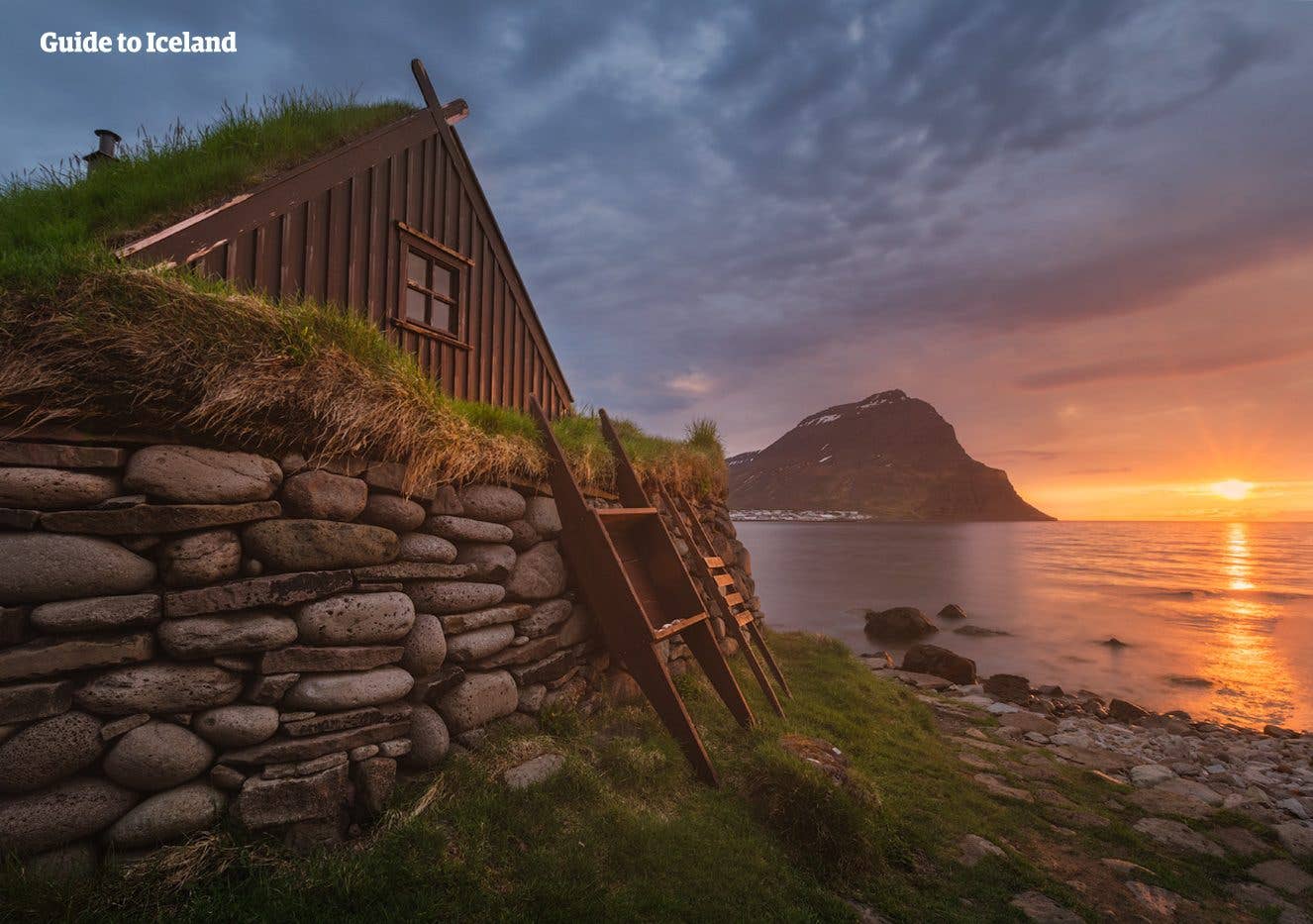 A grass-roofed turf house sits by the fjord at sunset near Bolungarvík in the Westfjords of Iceland.