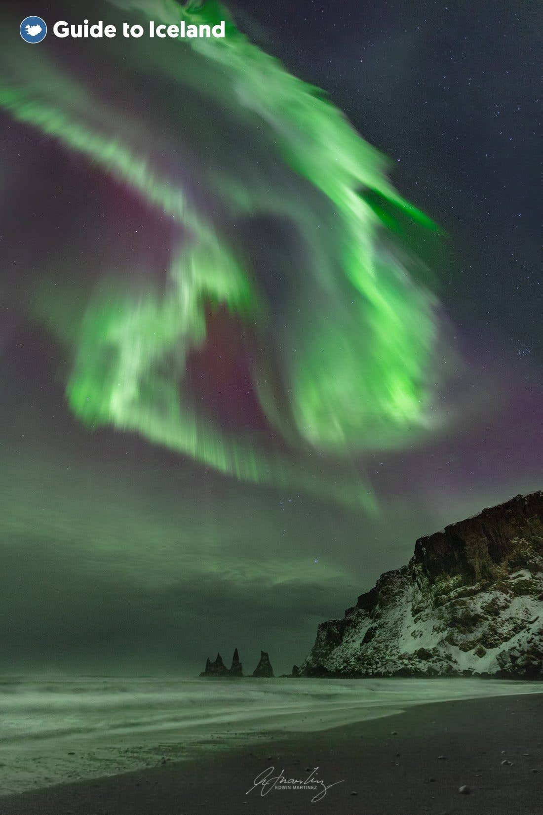 Green auroras dance above the cliffs and black beach at Reynisfjara near Vík, South Iceland.