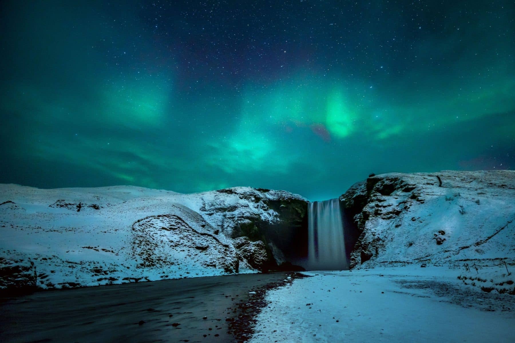 Green auroras fill the winter sky over the snow-framed Skógafoss waterfall in South Iceland.