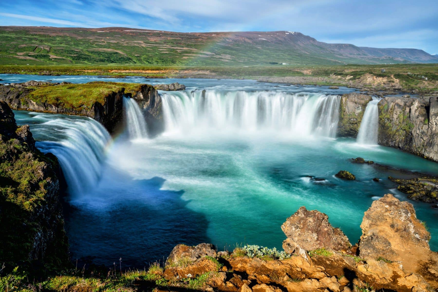 Turquoise water sweeps over the curved Goðafoss waterfall on a bright day in North Iceland.
