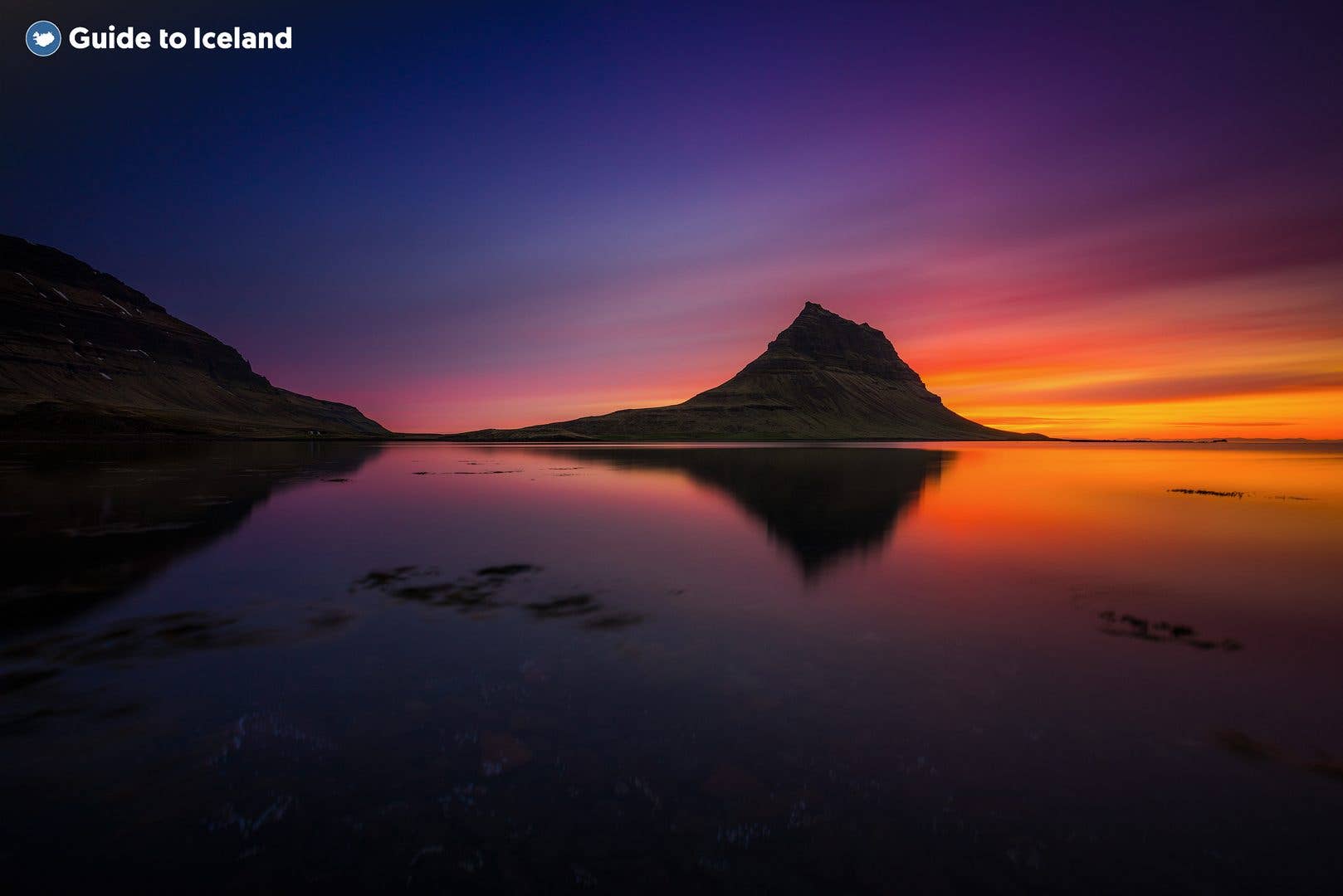 The setting sun silhouettes Kirkjufell mountain above a mirror-calm lagoon in West Iceland.