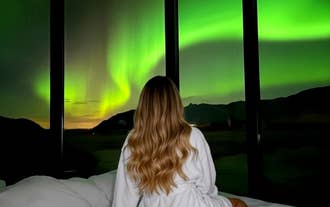 Woman in a robe sits on the bed watching the northern lights through the glass wall of a cabin in South Iceland.