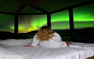 Woman in a robe sits on the bed watching the northern lights through the glass wall of a cabin in South Iceland.