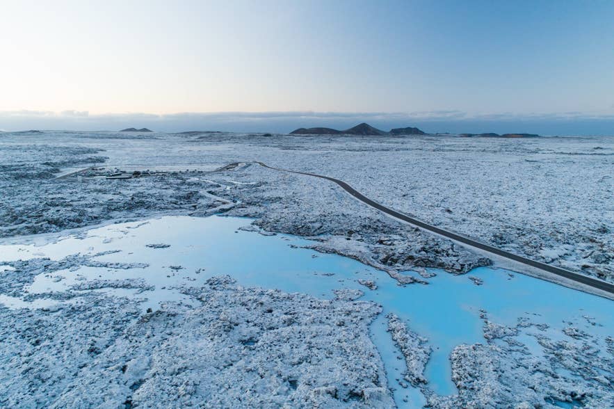 Aerial view of the Blue Lagoon in winter, its milky blue geothermal water winding through snow-covered lava fields on the Reykjanes peninsula in Iceland, with the spa buildings and access road in the distance at dusk
